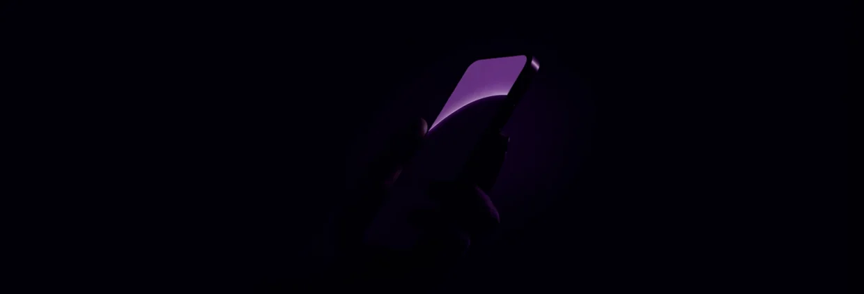 Mão segurando um celular com a tela de uma loja aberta, o preço e o botão de comprar em destaque, sobre fundo escuro com luz roxa, representando a compra que acontece na tela pequena.